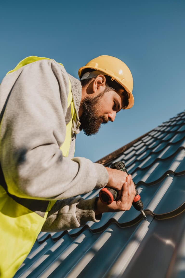 Travailleur en gilet de sécurité jaune et casque, utilisant une perceuse pour fixer des tôles métalliques sur un toit sous un ciel dégagé.